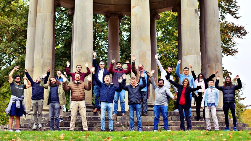 A group of persons facing the camera is standing in front of what appears to be a temple-like circular building with grey stone columns. All persons have raised their hands and are waving.