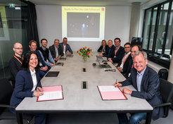 Photo of 12 persons at a long table, six on the right and six on the left. The two people at the front are signing a document and smile at the camera.